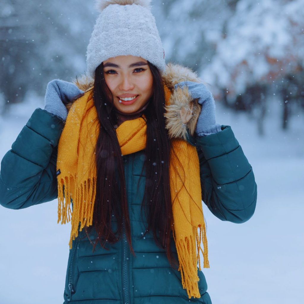 asian woman outside in the snow wearing a beanie hat, a yellow scarf, a green coat, and gloves. 